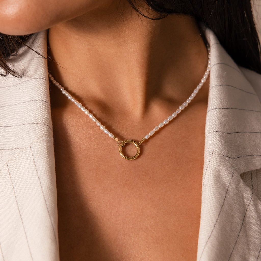 Close-up of a woman wearing the Pearl Chain Clasp Necklace in 18K Gold with a gold circular pendant, styled with a cream pinstripe blazer—a timeless piece from our collection.