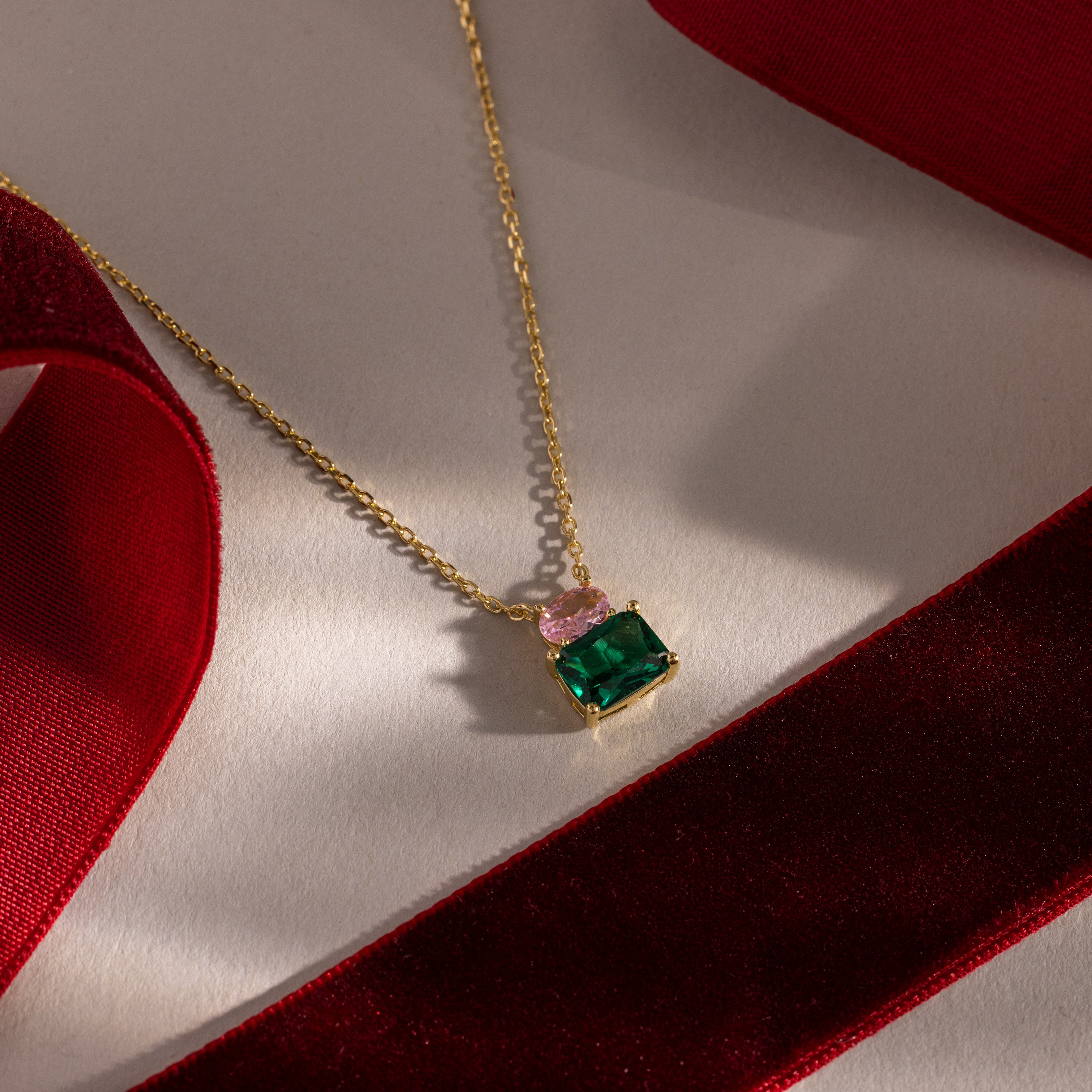 Flatlay of our Emerald Bloom Necklace with Pink Tourmaline and Emerald Gemstones against red ribbons