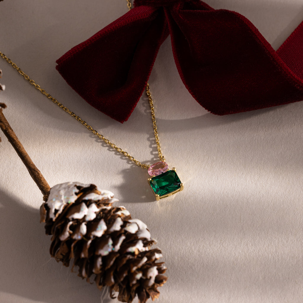 Flatlay of our Emerald Bloom Necklace with Pink Tourmaline and Emerald Gemstones against red ribbons and a pinecone