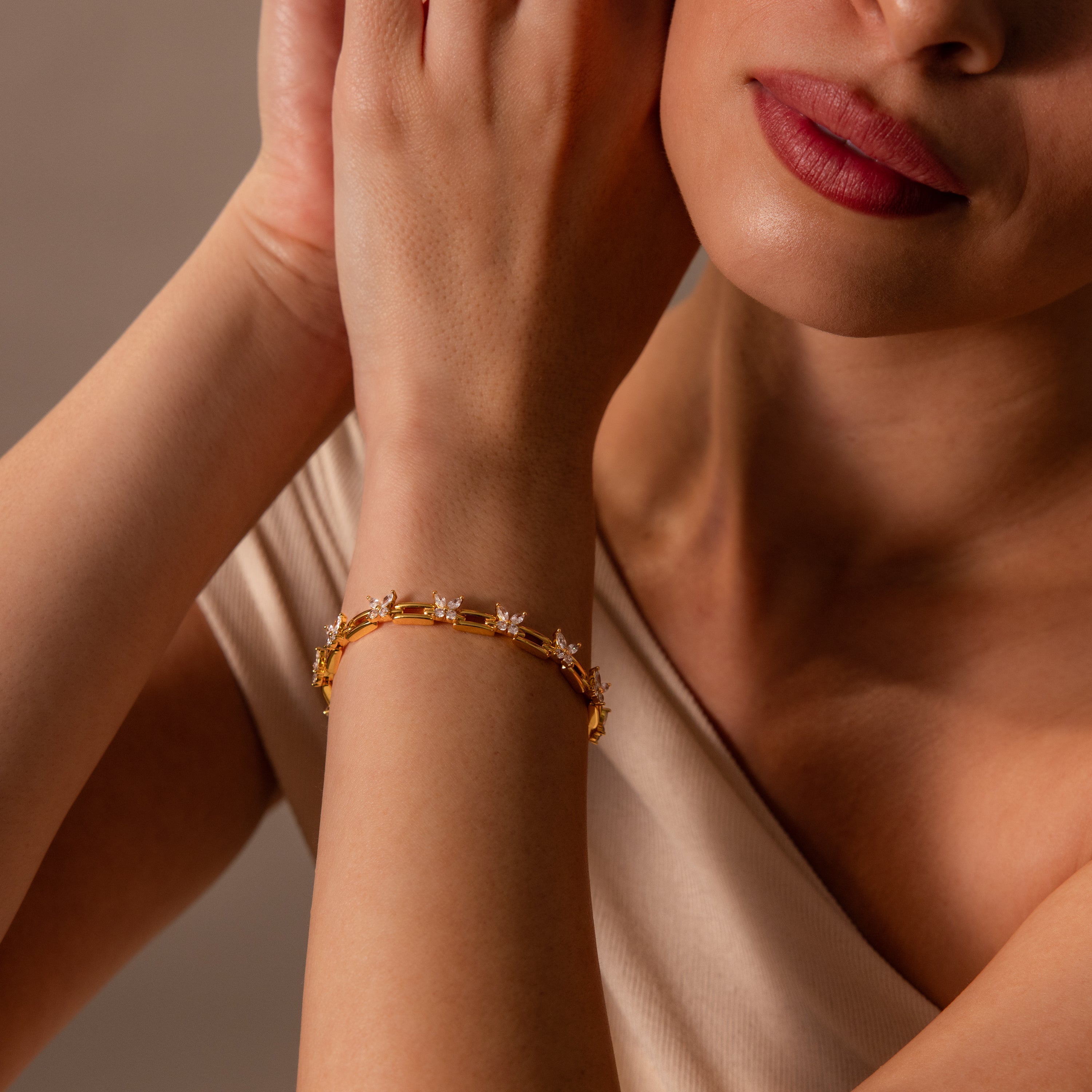 A woman with red lipstick gently touches her face, wearing the Mariposa Link Bracelet—a gold butterfly design with floral details that's the perfect feminine jewelry piece.