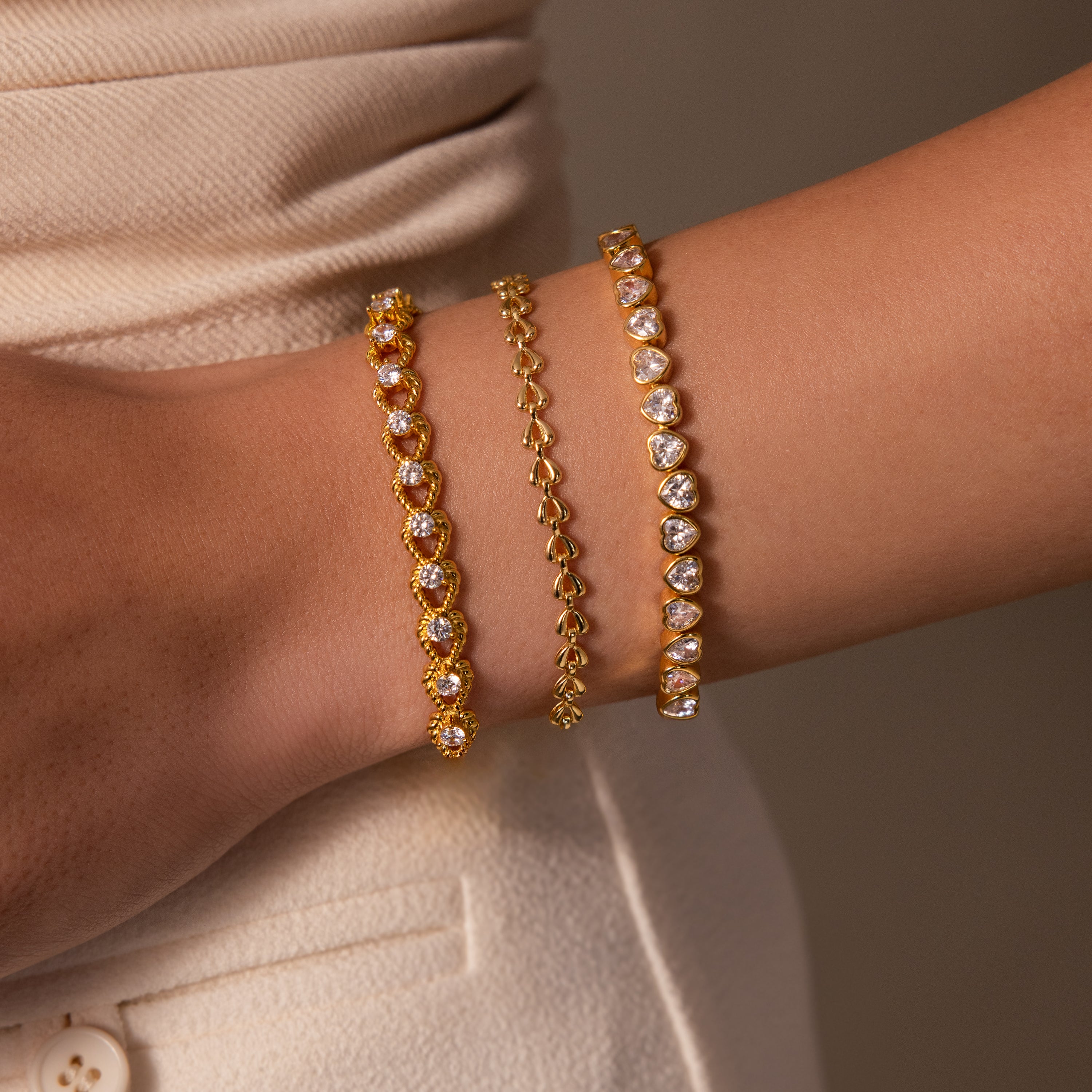 The wrist is adorned with the Diamond Heart Rope Bracelet, featuring sparkling diamond hearts, styled with cream-colored clothing.