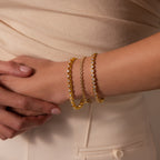 A woman in beige clothing wears elegant jewelry, showcasing the Heart Tennis Bracelet in 18K Gold as she rests her hand on her hip.