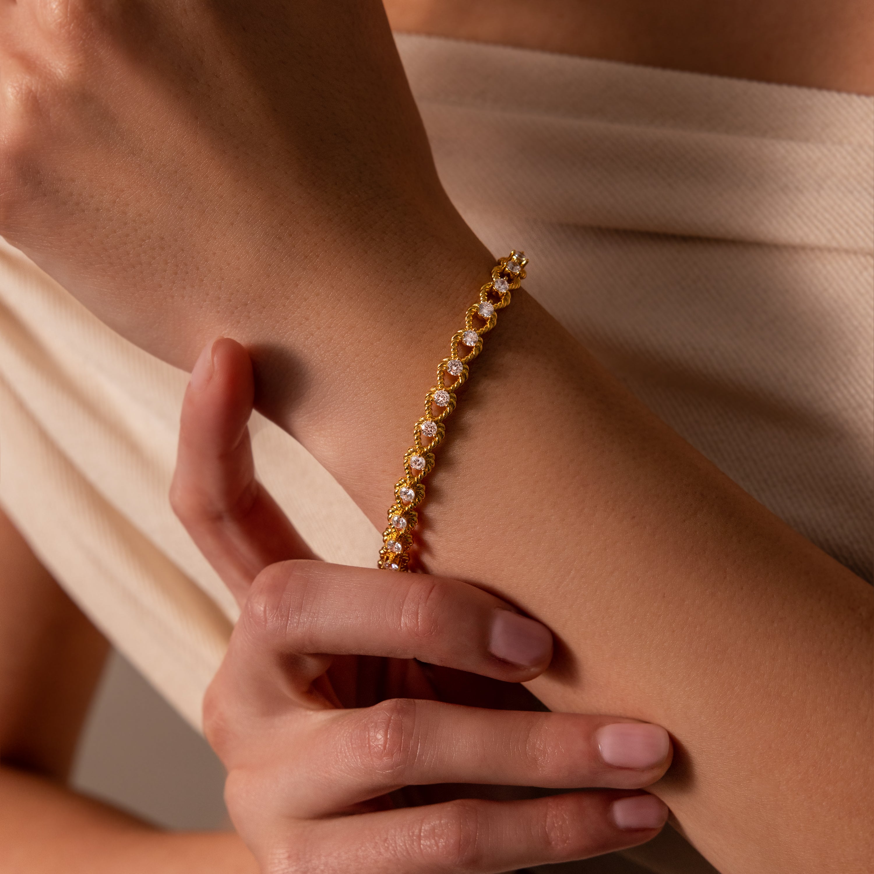 A woman’s hand wears the Diamond Heart Rope Bracelet, featuring clear gemstones and gold, set against a beige textured fabric background.