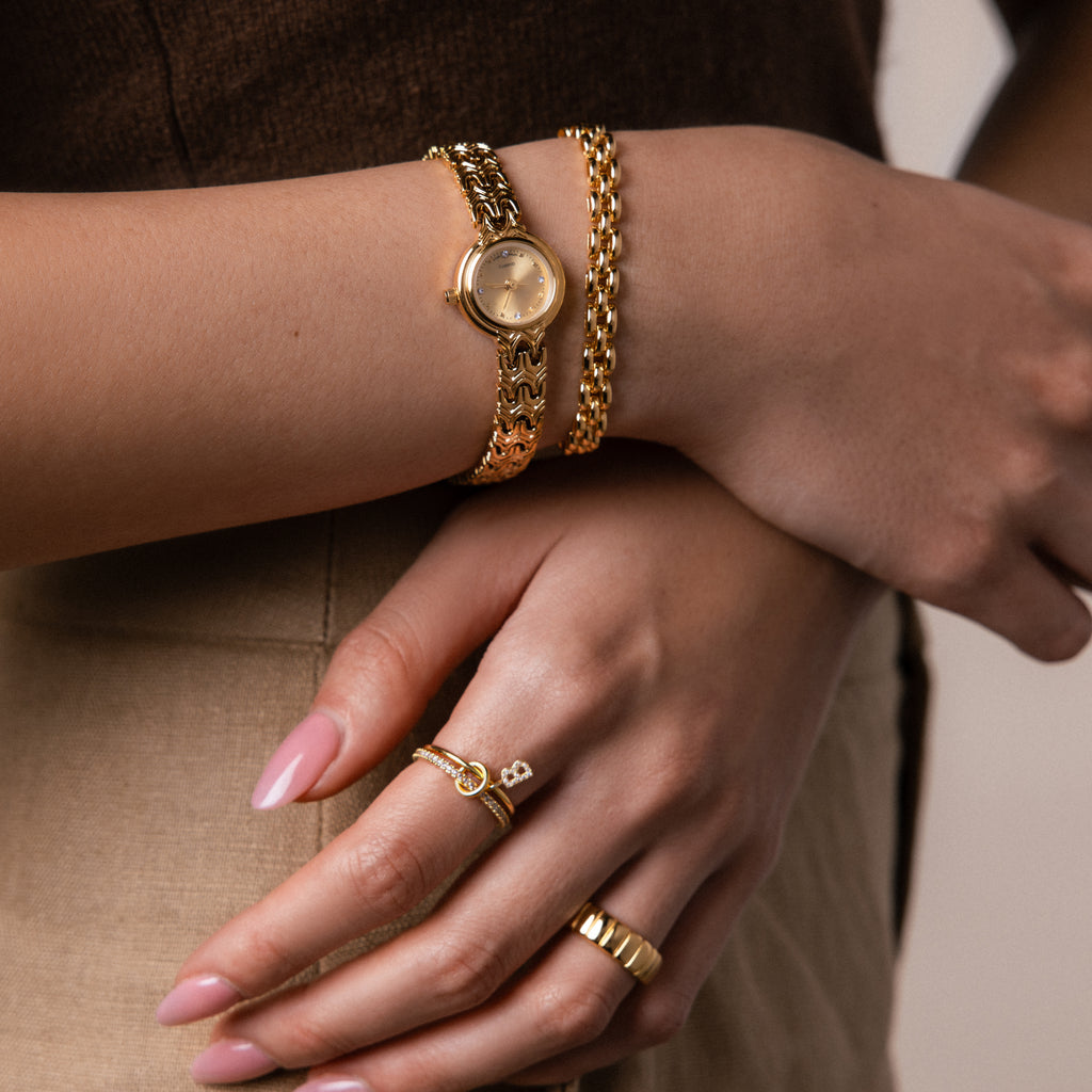 Close-up of a model's wrist and hand wearing gold jewelry look featuring our Wavy Link Timepiece watch, strap bracelet, Audrey ring and birthstone initial linked ring set.