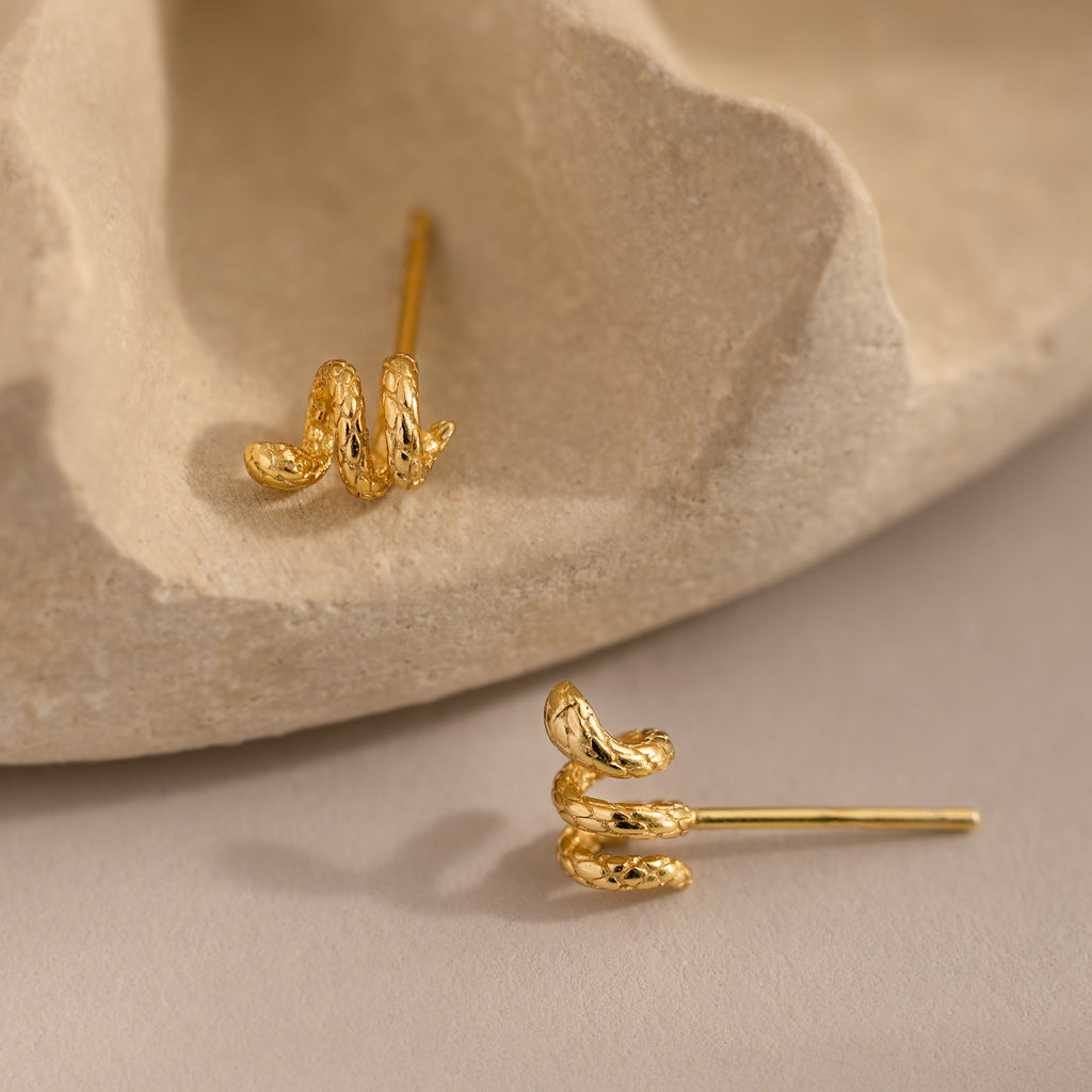 Gold 3D twisted snake stud earrings resting against a neutral background