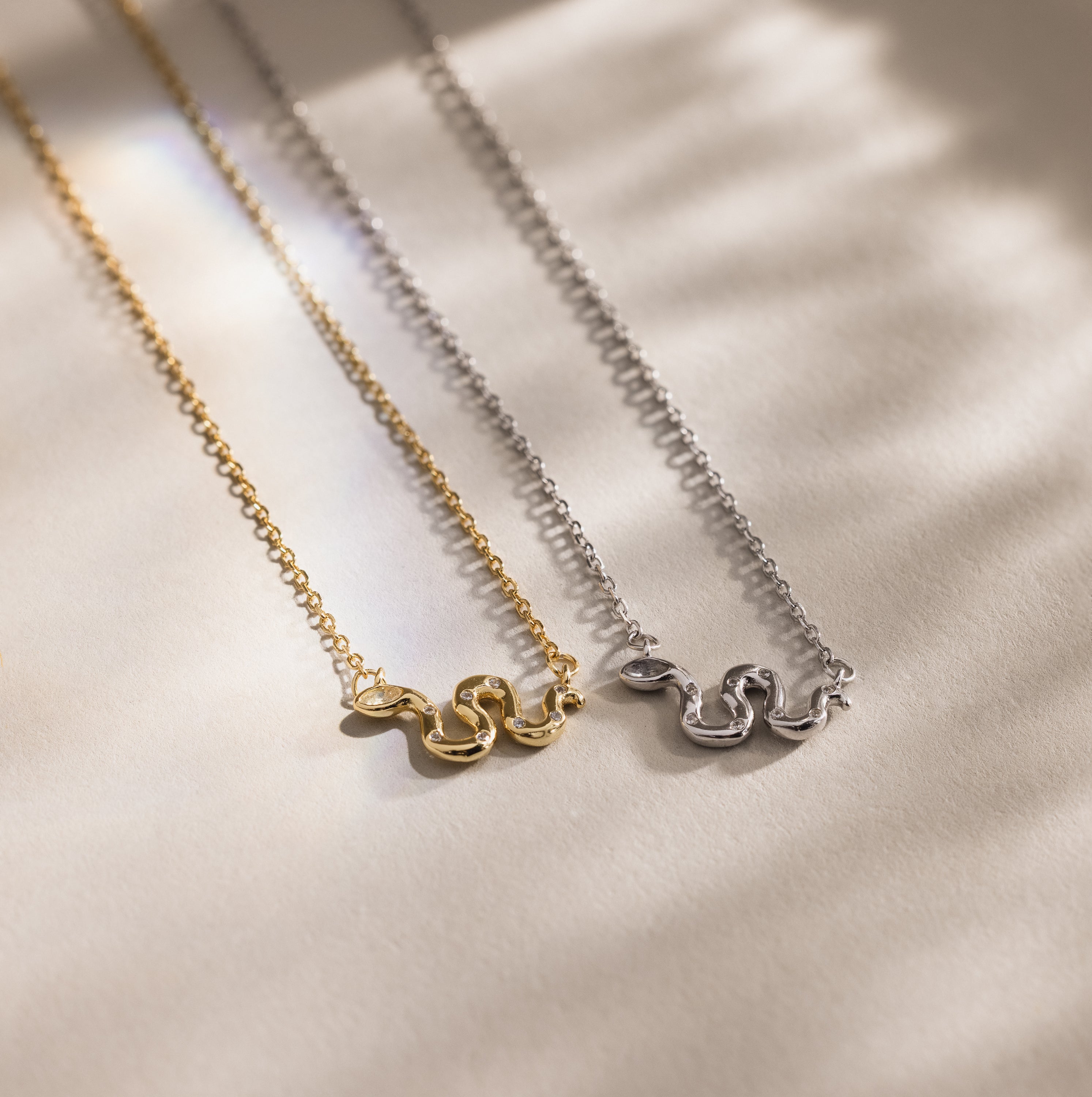 Gold and silver snake charm necklaces with pavé details, displayed side by side on a neutral background.