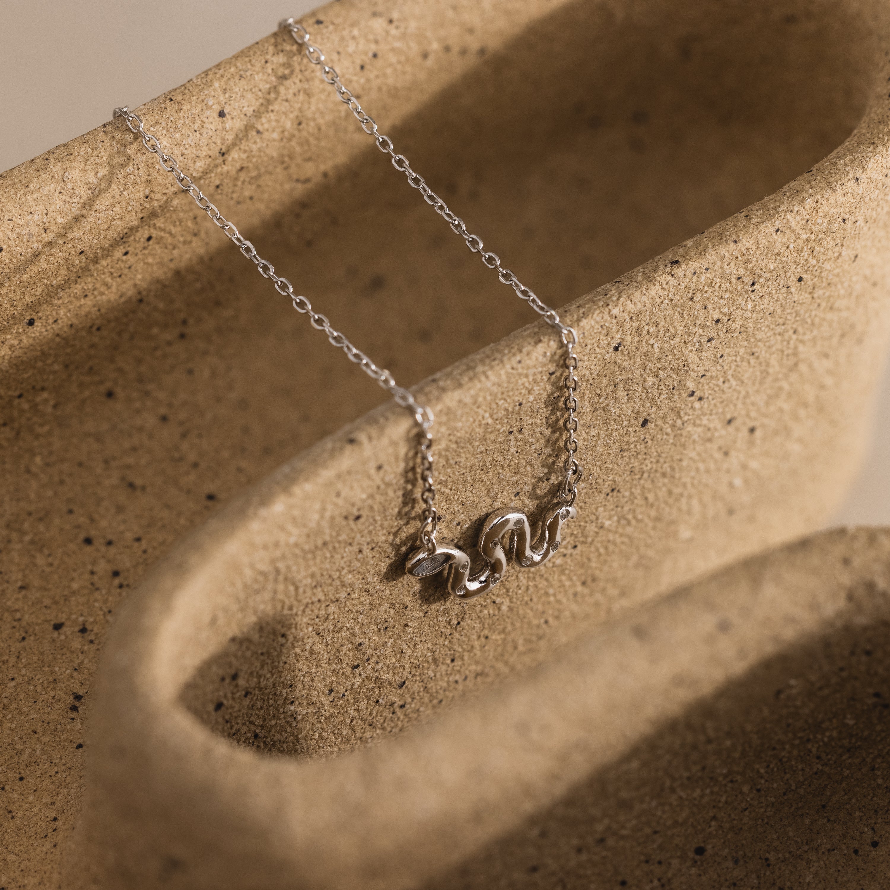 Close-up of a silver snake charm necklace displayed on a textured ceramic surface.