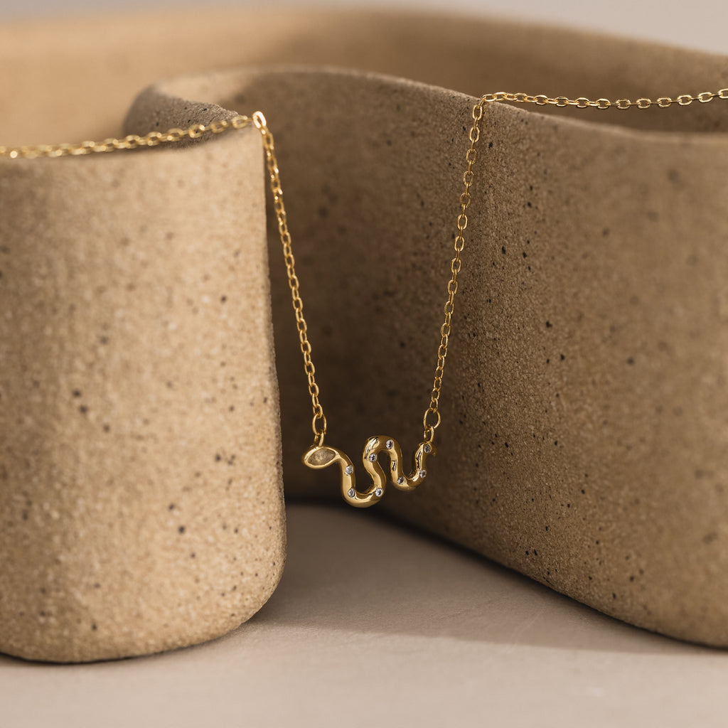 Close-up of a gold viper snake pendant necklace with pavé accents, displayed against a textured ceramic background.