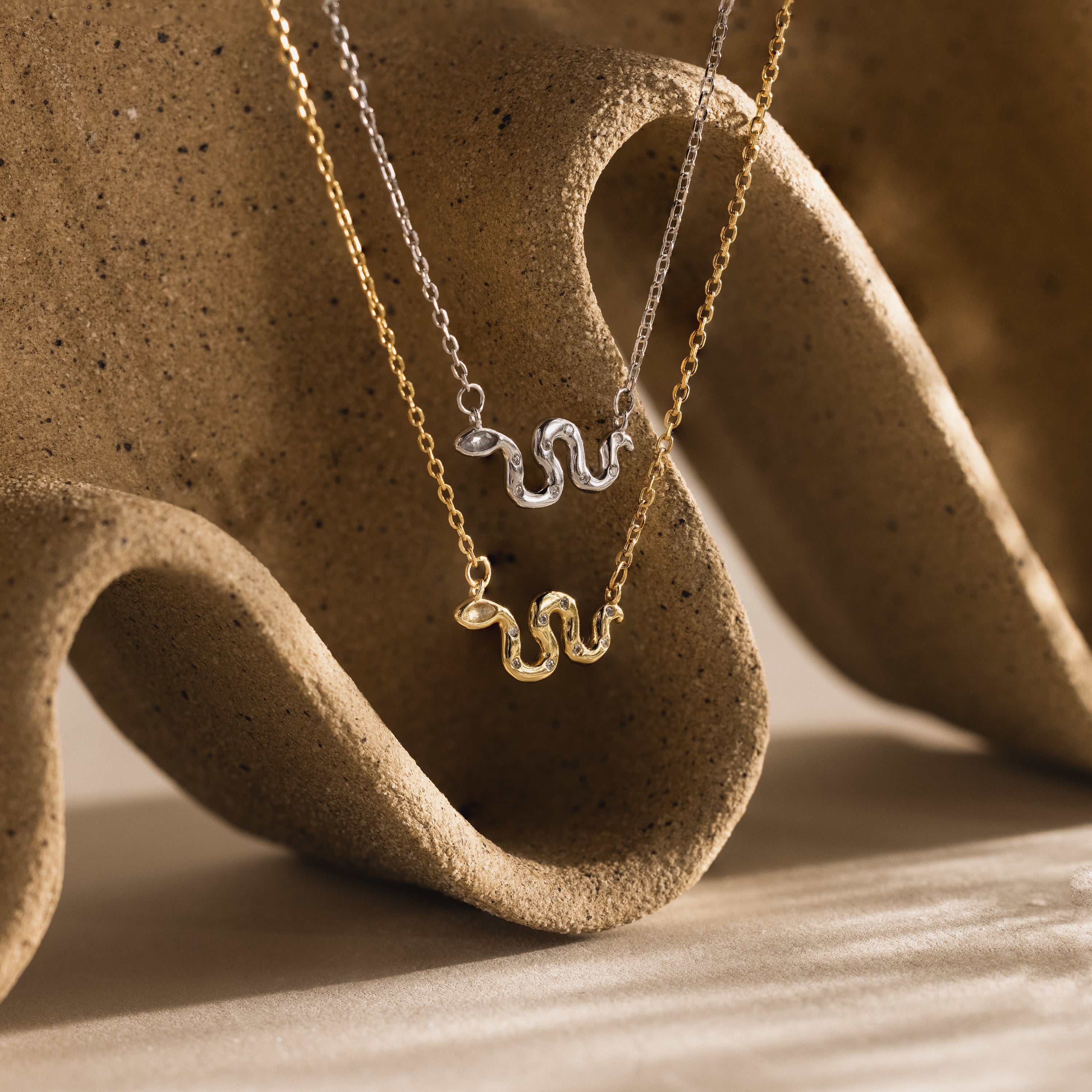 Gold and silver viper snake charm necklaces displayed over a sculpted ceramic surface with warm lighting.