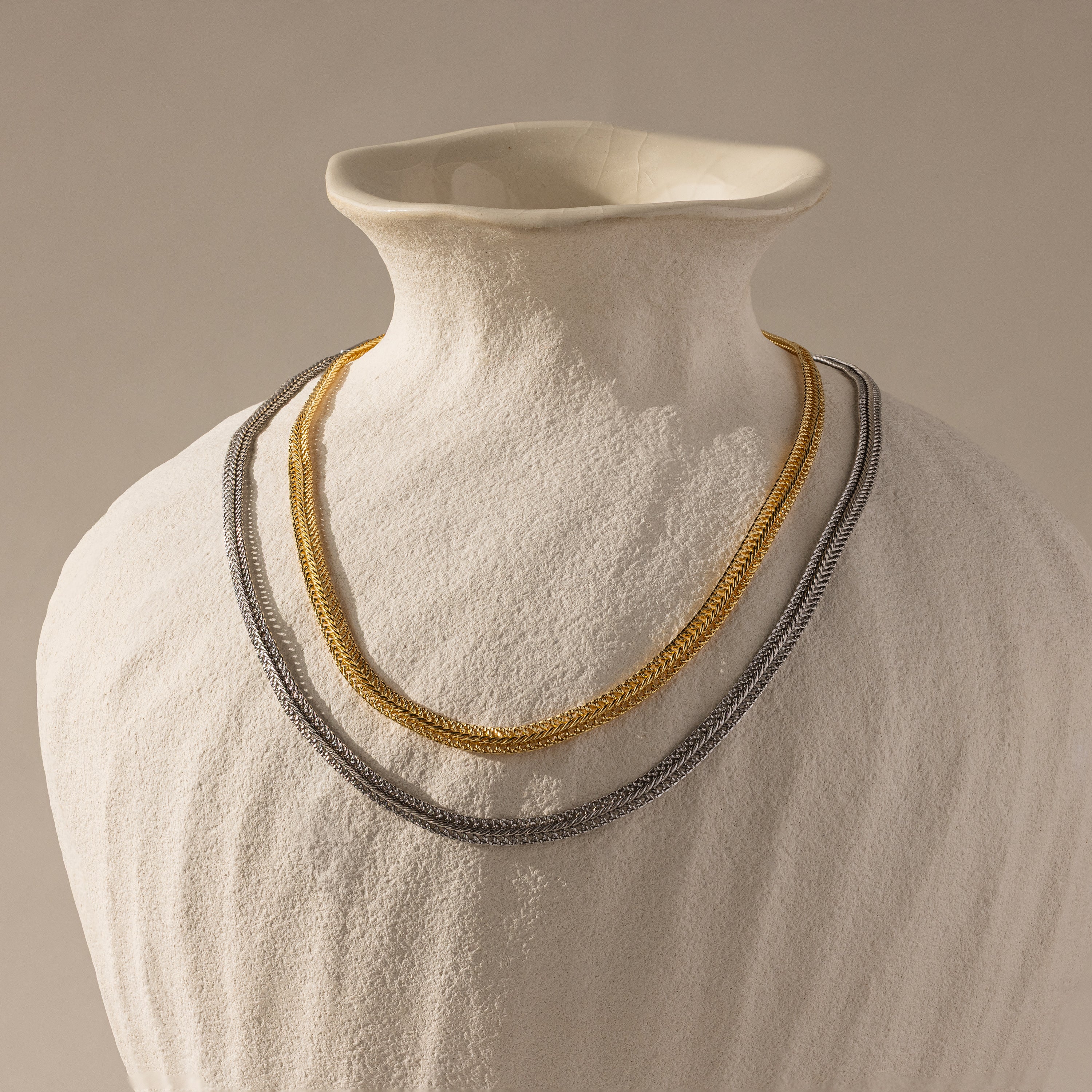 A Thin Mesh Chain Necklace in 18K Gold is draped on a white ceramic vase against a neutral background.