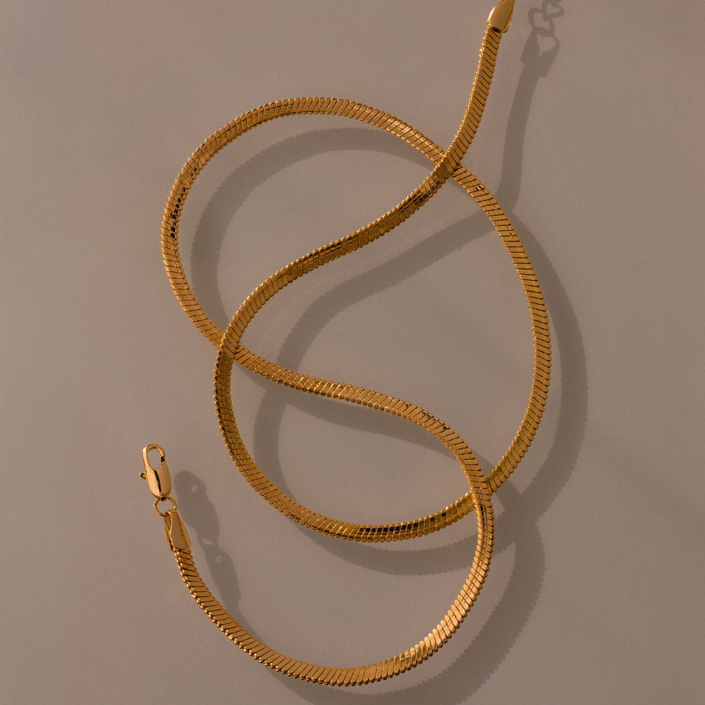The Twisted Snake Chain Necklace in 18K Gold, featuring a lobster clasp and arranged in loose curves on a neutral background, is perfect as modern jewelry and ideal for layering.