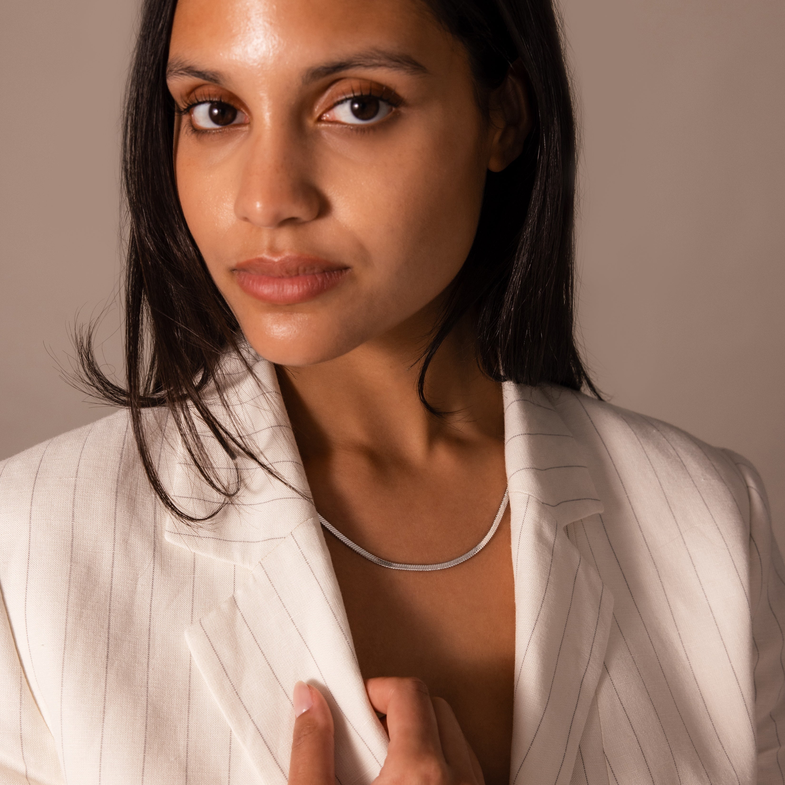 A woman wearing a white pinstripe blazer and the Twisted Snake Chain Necklace in Silver looks at the camera with a neutral expression.