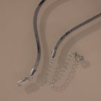 Modern jewelry at its finest—the Twisted Snake Chain Necklace in Silver features a smooth silver chain, clasp, and extender with an open heart charm, showcased on a beige background.