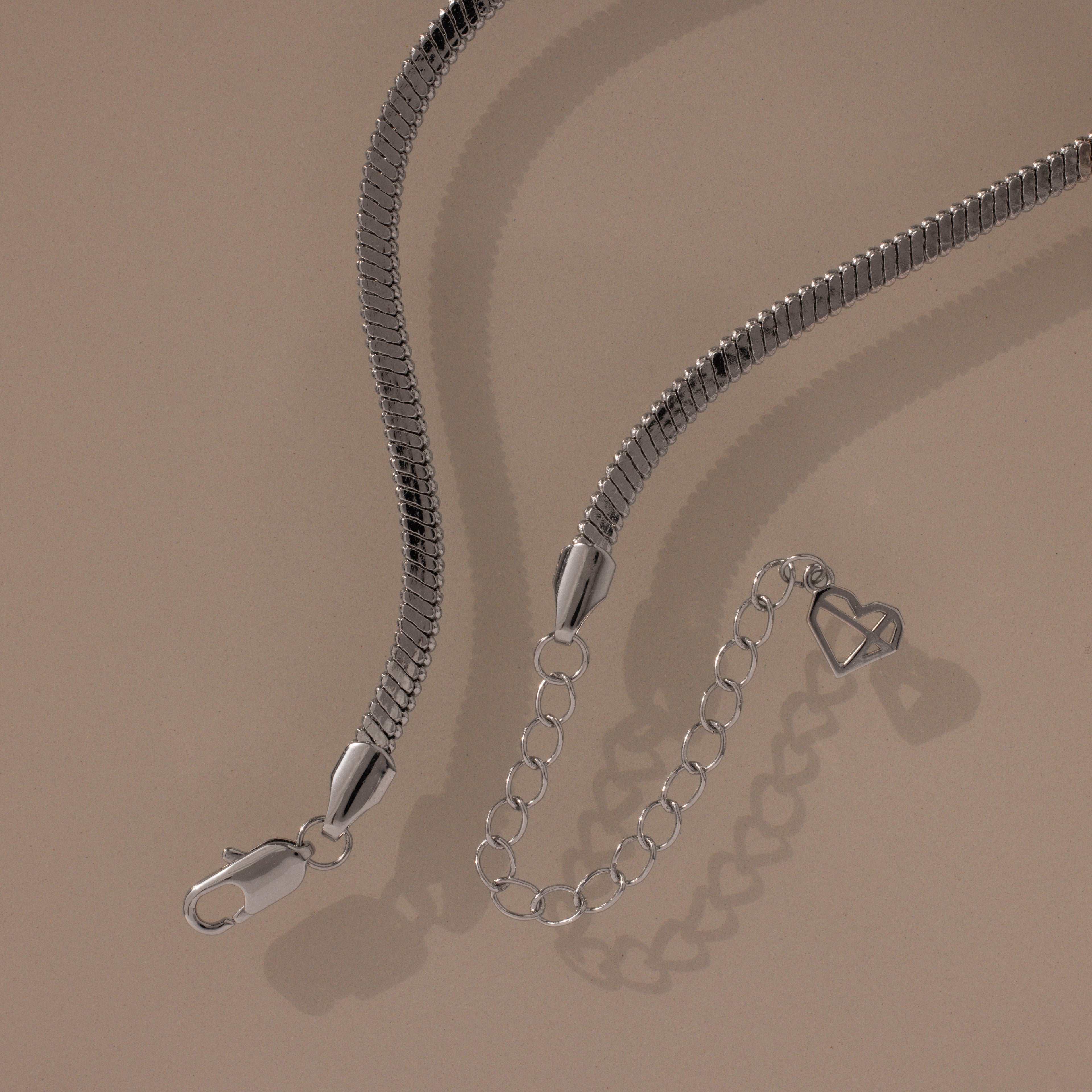 Modern jewelry at its finest—the Twisted Snake Chain Necklace in Silver features a smooth silver chain, clasp, and extender with an open heart charm, showcased on a beige background.