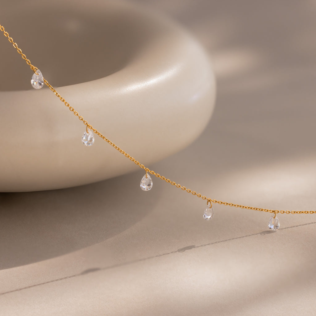Delicate gold chain necklace featuring evenly spaced teardrop-shaped diamond charms, gracefully draped in front of a smooth, rounded ceramic object on a soft beige surface.
