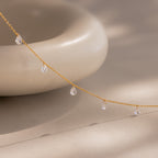 Delicate gold chain necklace featuring evenly spaced teardrop-shaped diamond charms, gracefully draped in front of a smooth, rounded ceramic object on a soft beige surface.