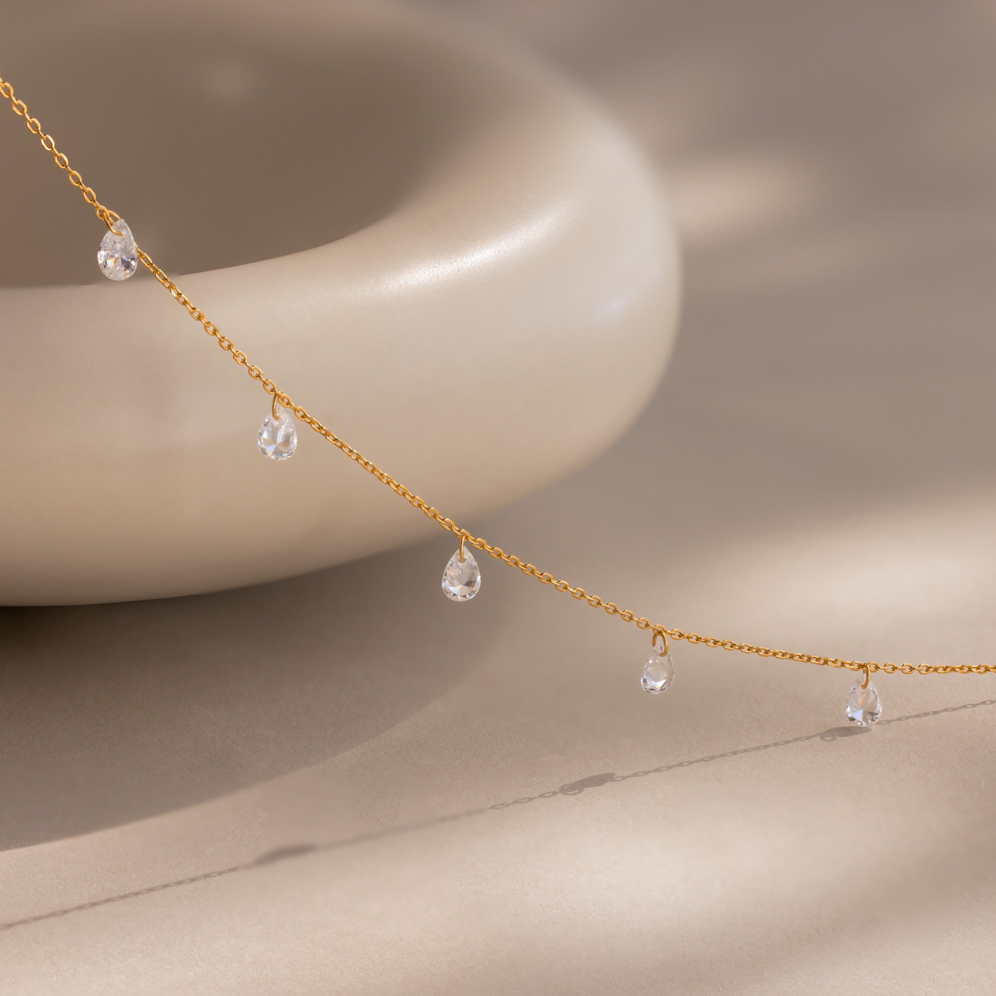 Delicate gold chain necklace featuring evenly spaced teardrop-shaped diamond charms, gracefully draped in front of a smooth, rounded ceramic object on a soft beige surface.