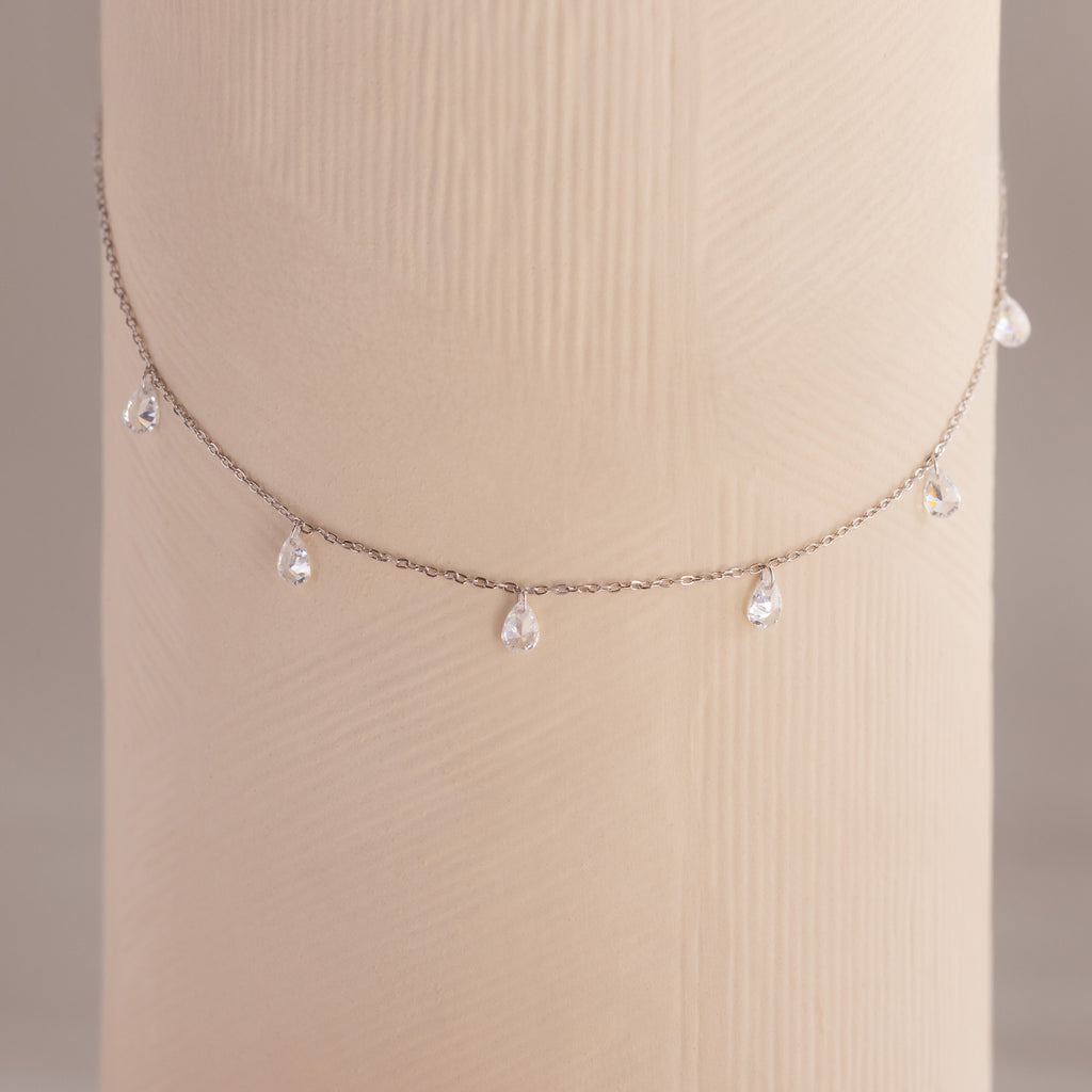 Diamond teardrop station necklace in silver draped around a textured cylindrical display in soft lighting.