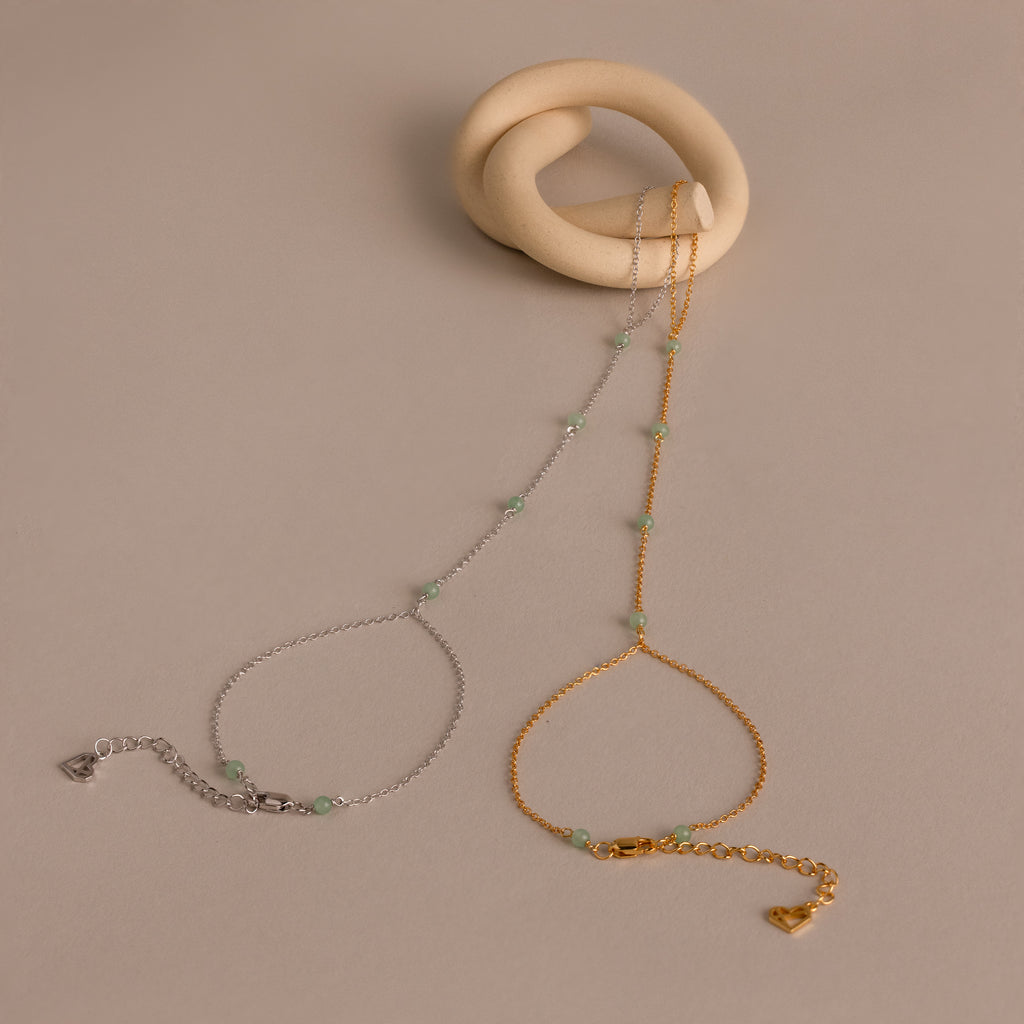 The Jade Hand Chain Bracelet is a delicate piece featuring small green jade beads and a heart clasp, available in gold or silver chains, shown on a beige background.