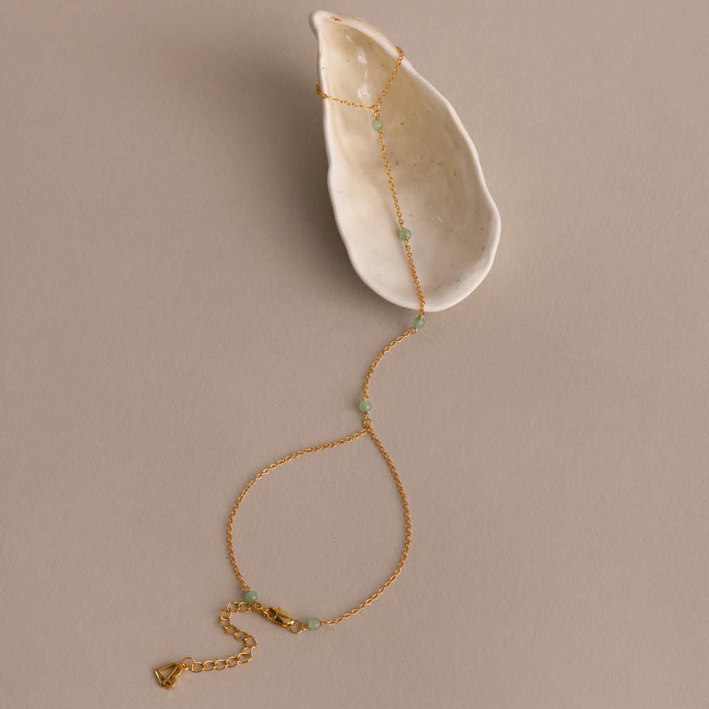 Jade Hand Chain Bracelet in 18K Gold with small jade beads displayed on a white ceramic dish against a beige background.