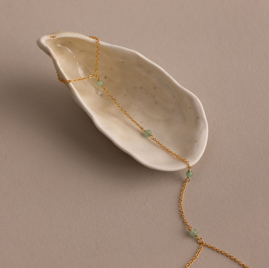The Jade Hand Chain Bracelet in 18K Gold is draped over a white ceramic shell on a beige surface, showcasing its delicate jade beads.