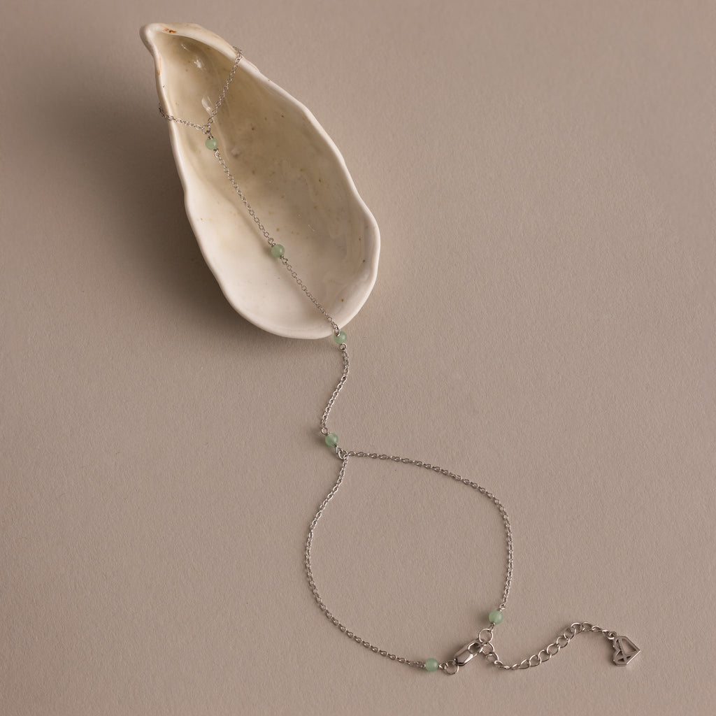 The Jade Hand Chain Bracelet in Silver is displayed on a cream-colored seashell, featuring a delicate silver chain adorned with jade beads.