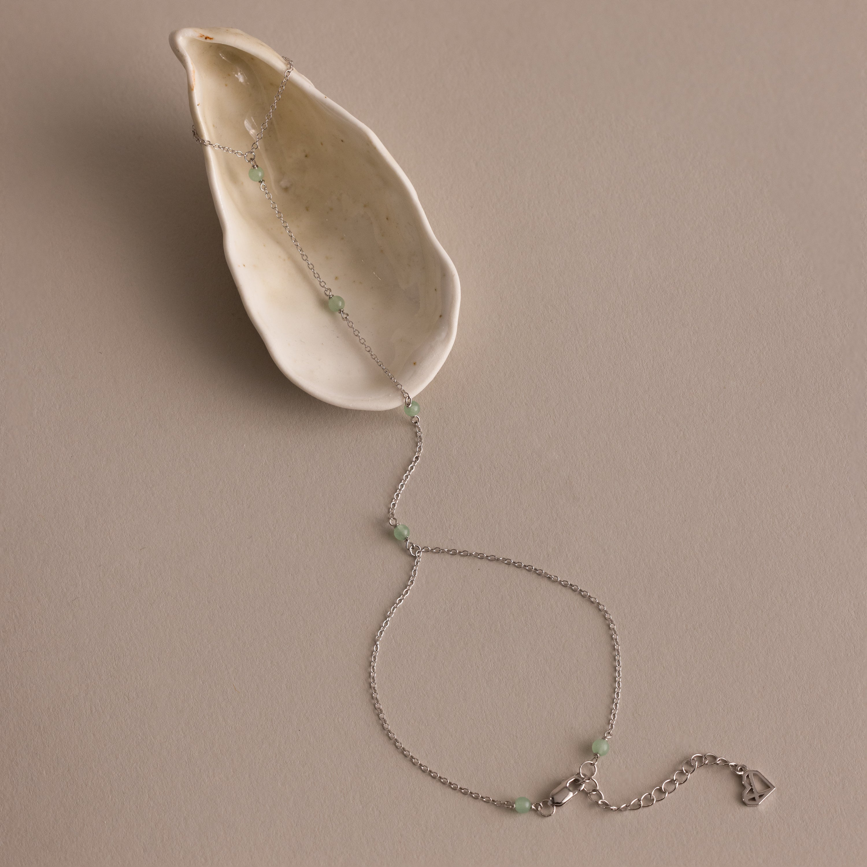 The Jade Hand Chain Bracelet features delicate silver and green jade beads, beautifully presented on a cream-colored shell.