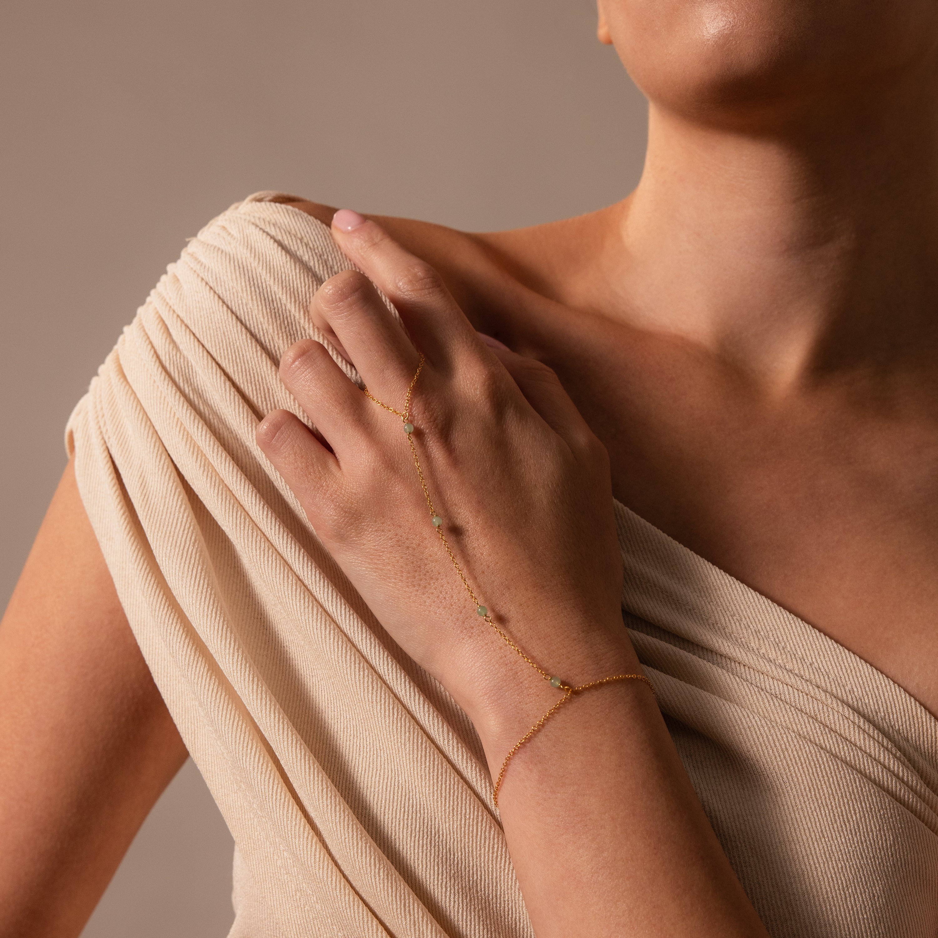 A woman wearing a beige dress and the Jade Hand Chain Bracelet in 18K Gold, adorned with delicate gemstones and jade beads.