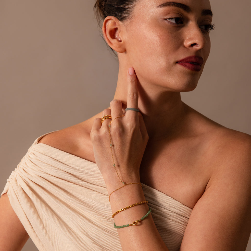 Woman in an off-shoulder beige top wears the Jade Hand Chain Bracelet in 18K Gold, featuring gold and green jade beads. She raises her hand to her face and looks sideways.
