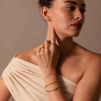 Woman in an off-shoulder beige top wears the Jade Hand Chain Bracelet in 18K Gold, featuring gold and green jade beads. She raises her hand to her face and looks sideways.