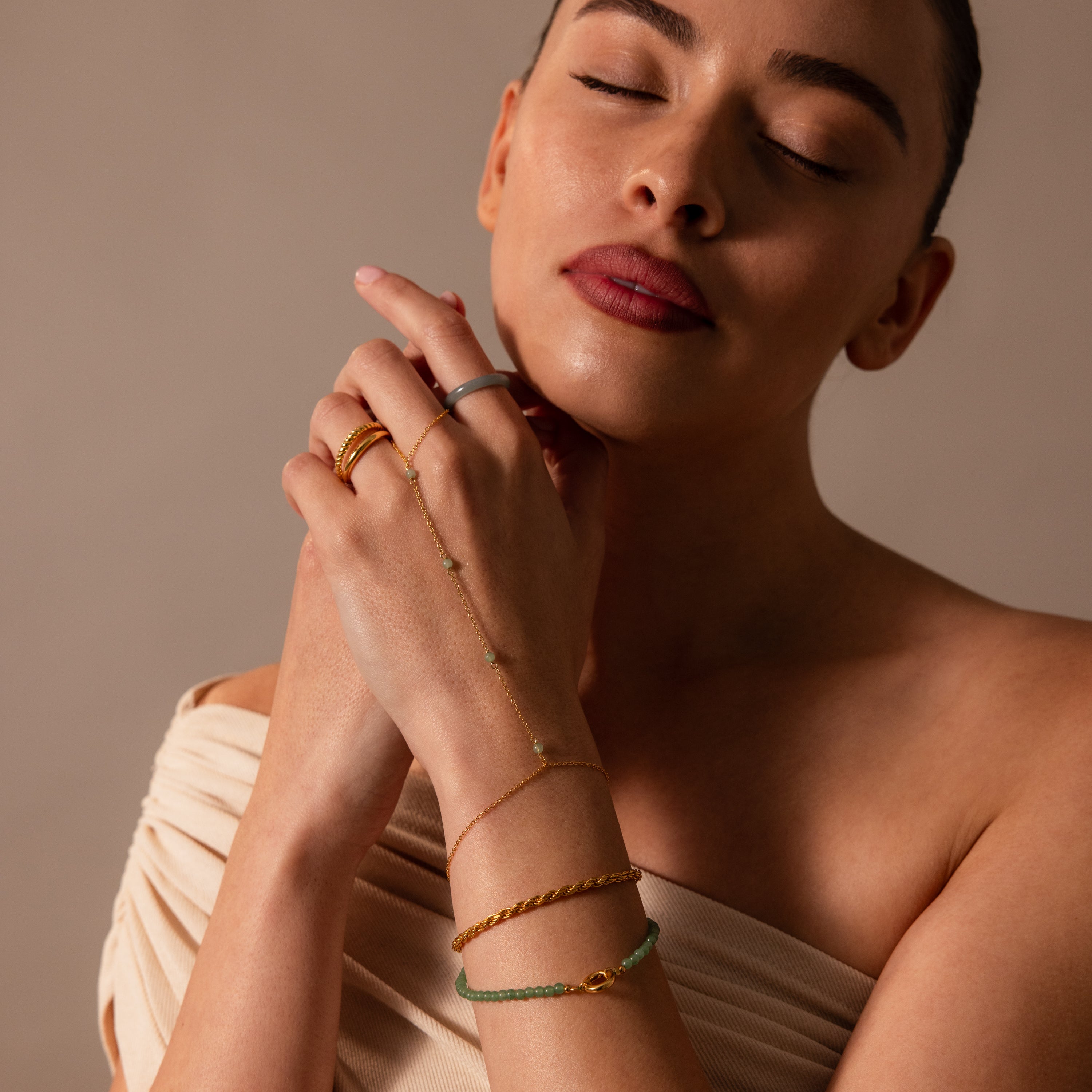 A woman with closed eyes poses gracefully with her hands near her face, showcasing the Jade Hand Chain Bracelet adorned with green jade beads and elegant gold jewelry.