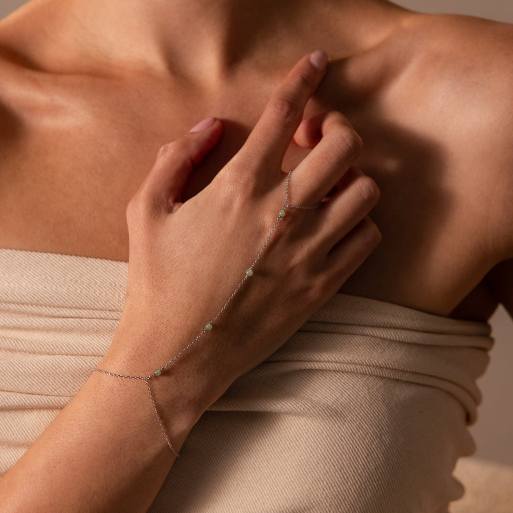 A hand adorned with the Jade Hand Chain Bracelet in Silver, featuring delicate beads, rests on a bare shoulder draped in cream-colored fabric.