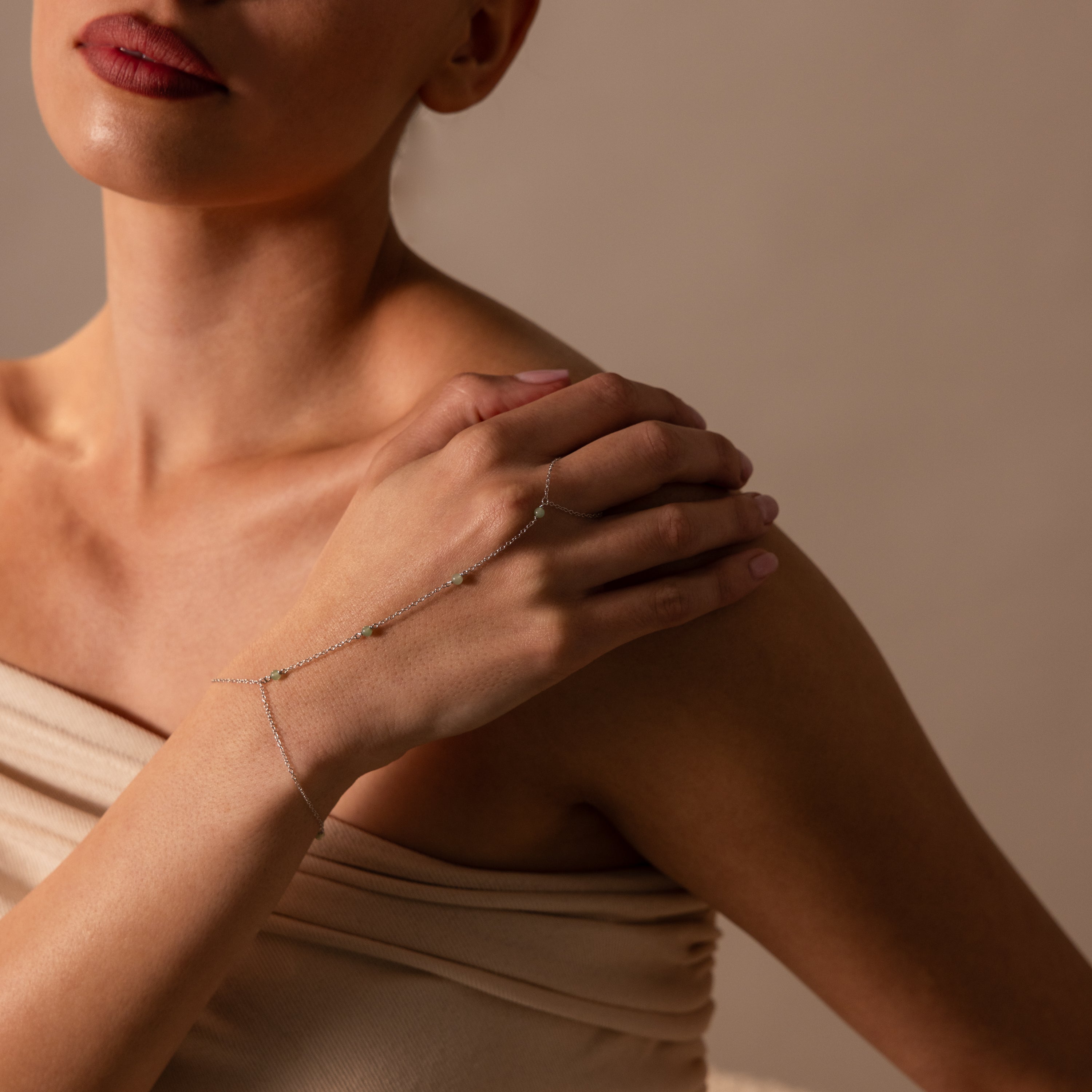 A woman in a beige top touches her shoulder, highlighting the Jade Hand Chain Bracelet with delicate green jade beads.