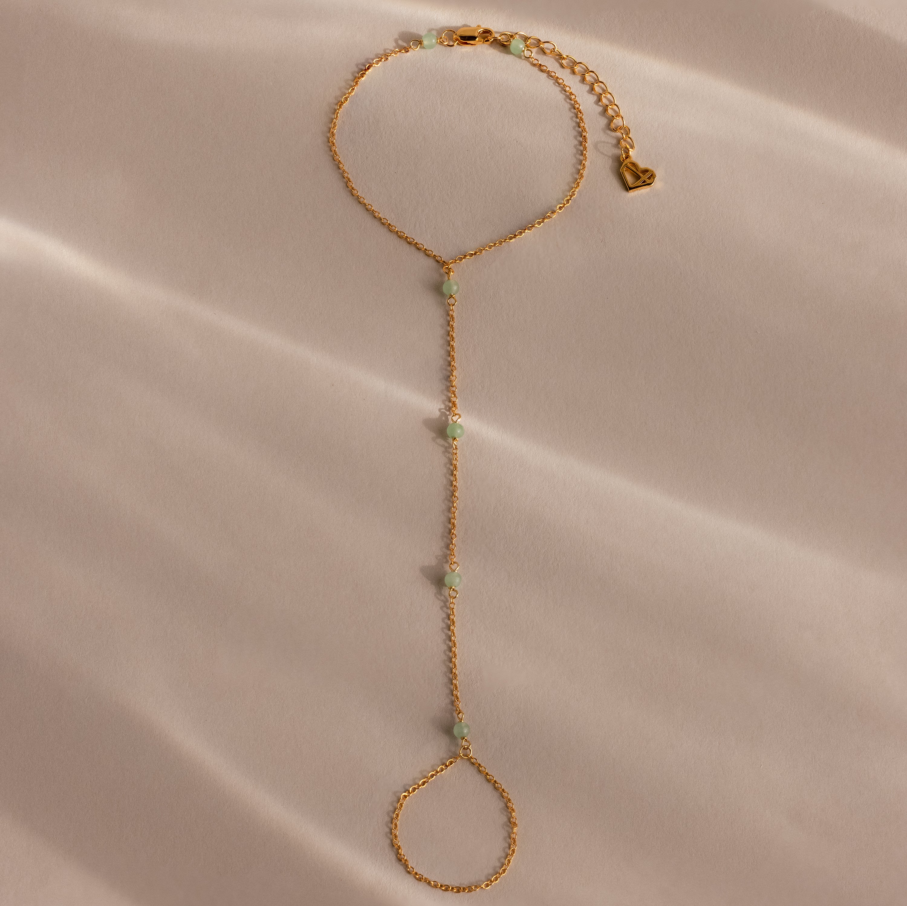 The Jade Hand Chain Bracelet features delicate green jade beads and is shown laid flat on a soft beige fabric background.