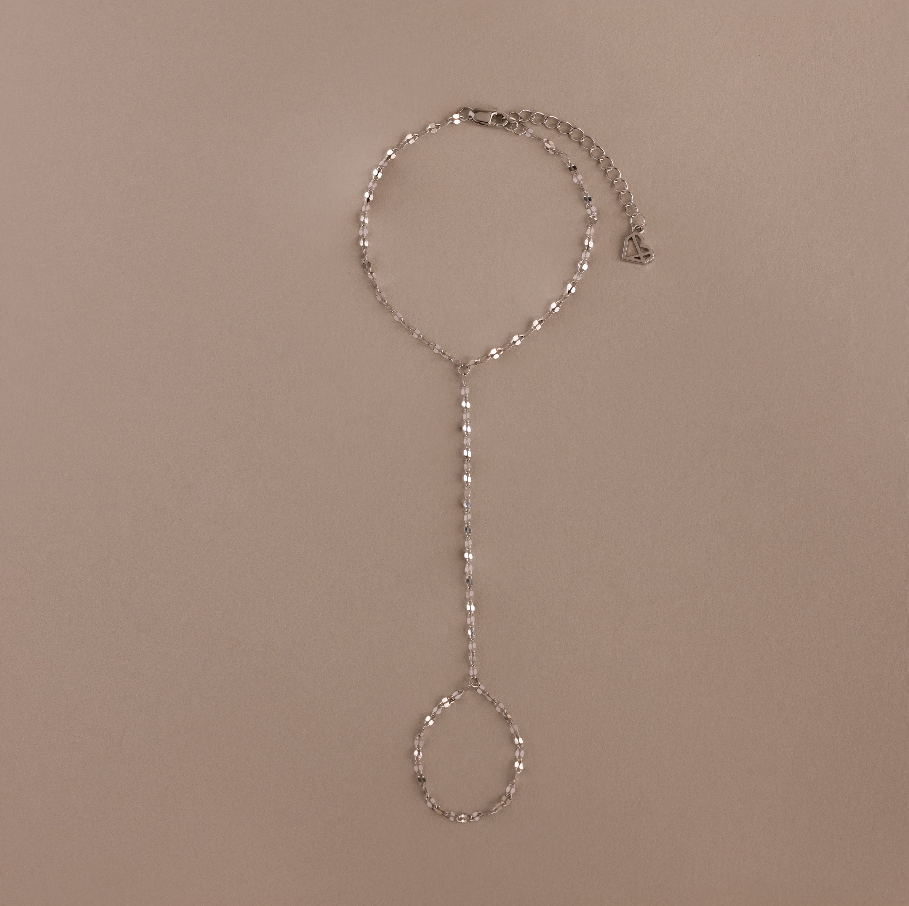 The Sparkle Hand Chain Bracelet in Silver features mirror cut links, a linked finger loop, and an adjustable clasp, beautifully displayed on a beige background.