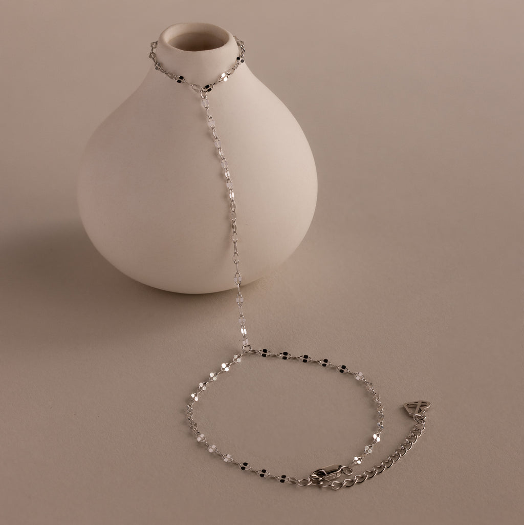 The Sparkle Hand Chain Bracelet in Silver, featuring mirror-cut links and black and clear beads, is draped over a round white vase on a neutral surface.
