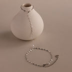 The Sparkle Hand Chain Bracelet in Silver, featuring mirror-cut links and black and clear beads, is draped over a round white vase on a neutral surface.