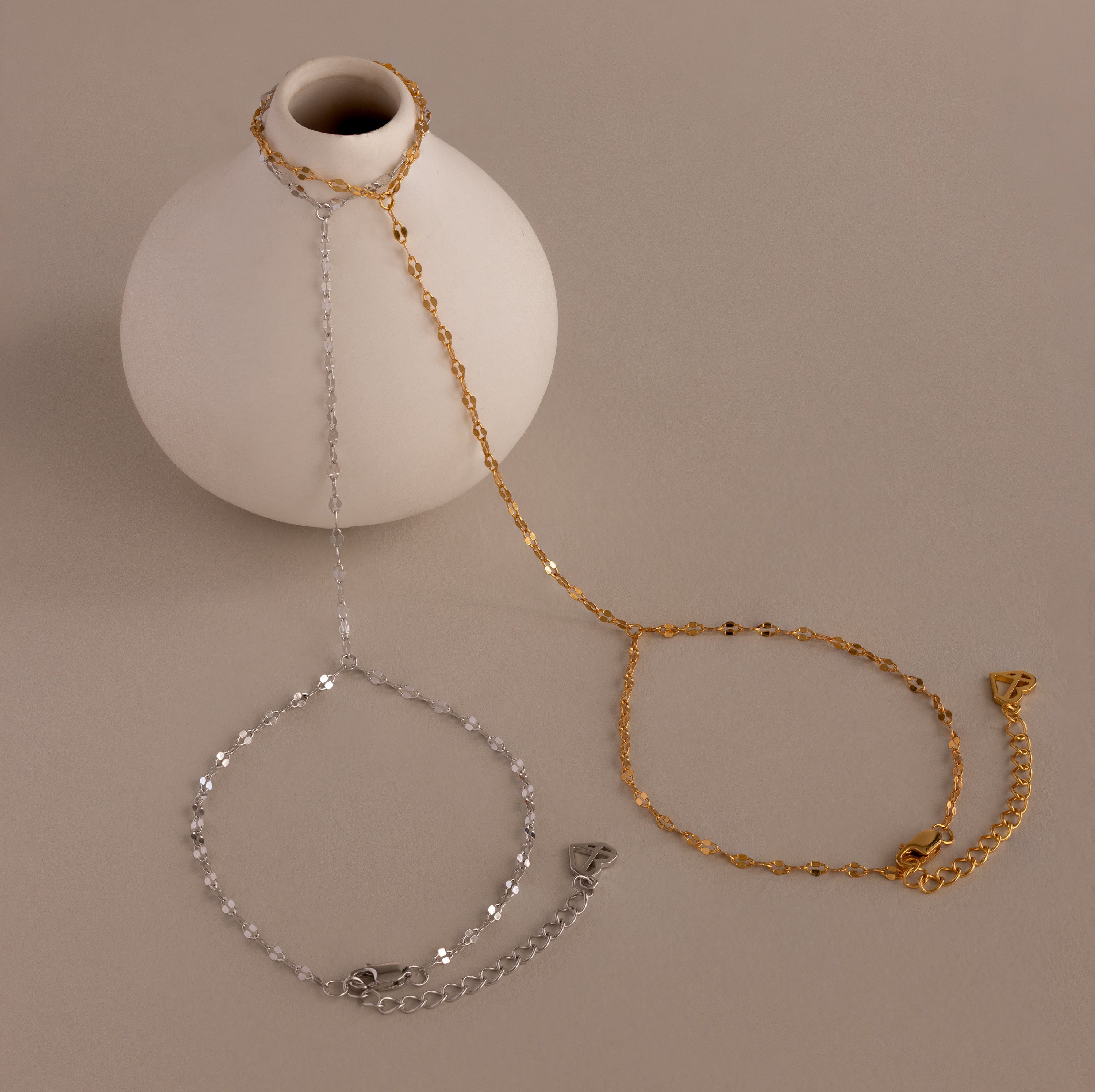 A white vase with Sparkle Hand Chain Bracelets, featuring gold and silver mirror-cut links, draped around it on a beige surface.