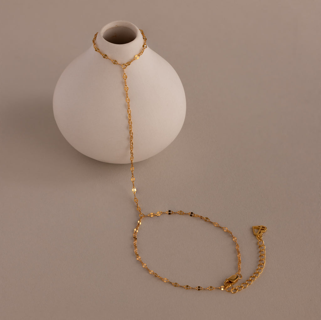 The Sparkle Hand Chain Bracelet in 18K Gold, featuring delicate mirror-cut links, is draped over a small round white vase on a beige surface.