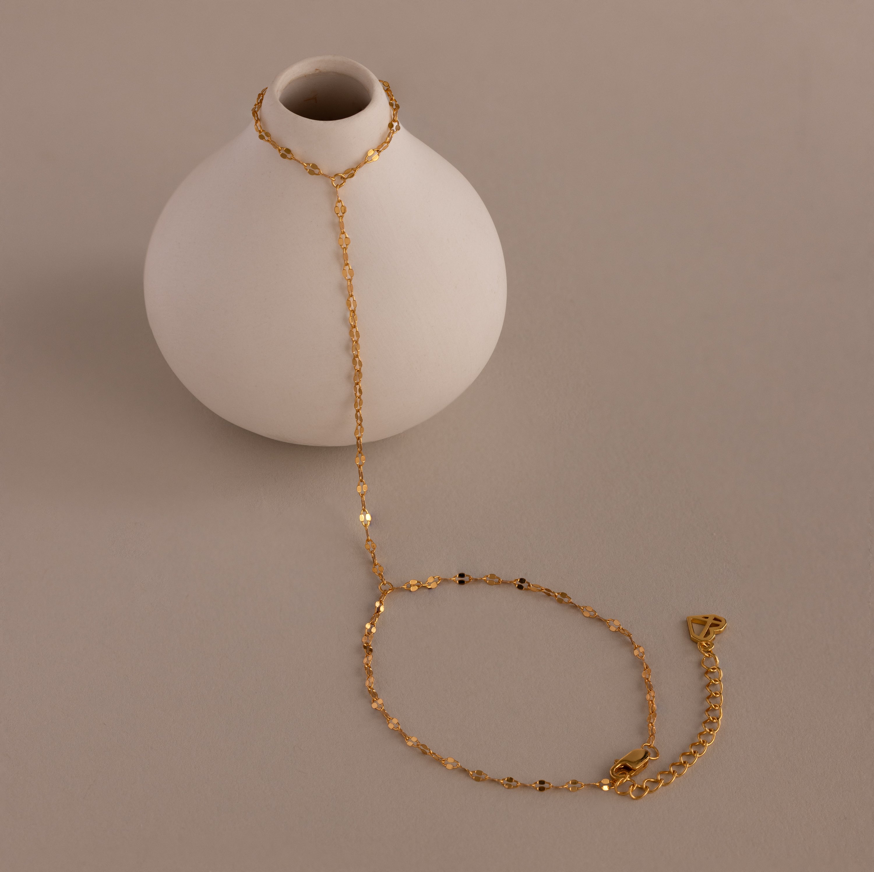 The Sparkle Hand Chain Bracelet in 18K Gold, featuring delicate mirror-cut links, is draped over a small round white vase on a beige surface.