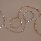 The Sparkle Hand Chain Bracelet features two delicate mirror-cut chains—one gold, one silver—arranged in loose loops on a neutral beige surface.