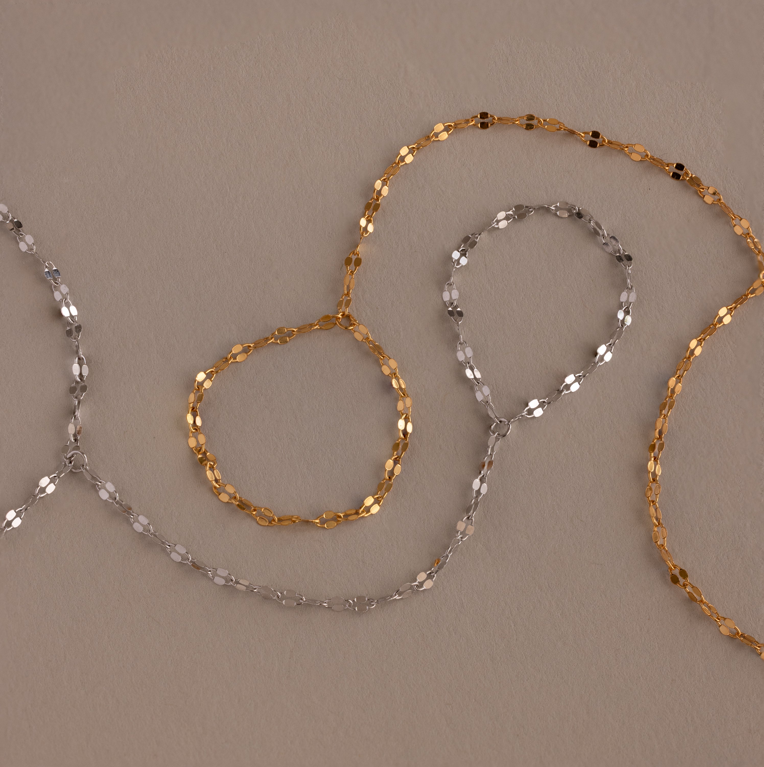 The Sparkle Hand Chain Bracelet features two delicate mirror-cut chains—one gold, one silver—arranged in loose loops on a neutral beige surface.