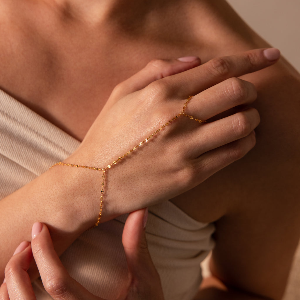 A woman’s hand adorned with the Sparkle Hand Chain Bracelet in 18K Gold, featuring delicate mirror cut links, resting on her chest.
