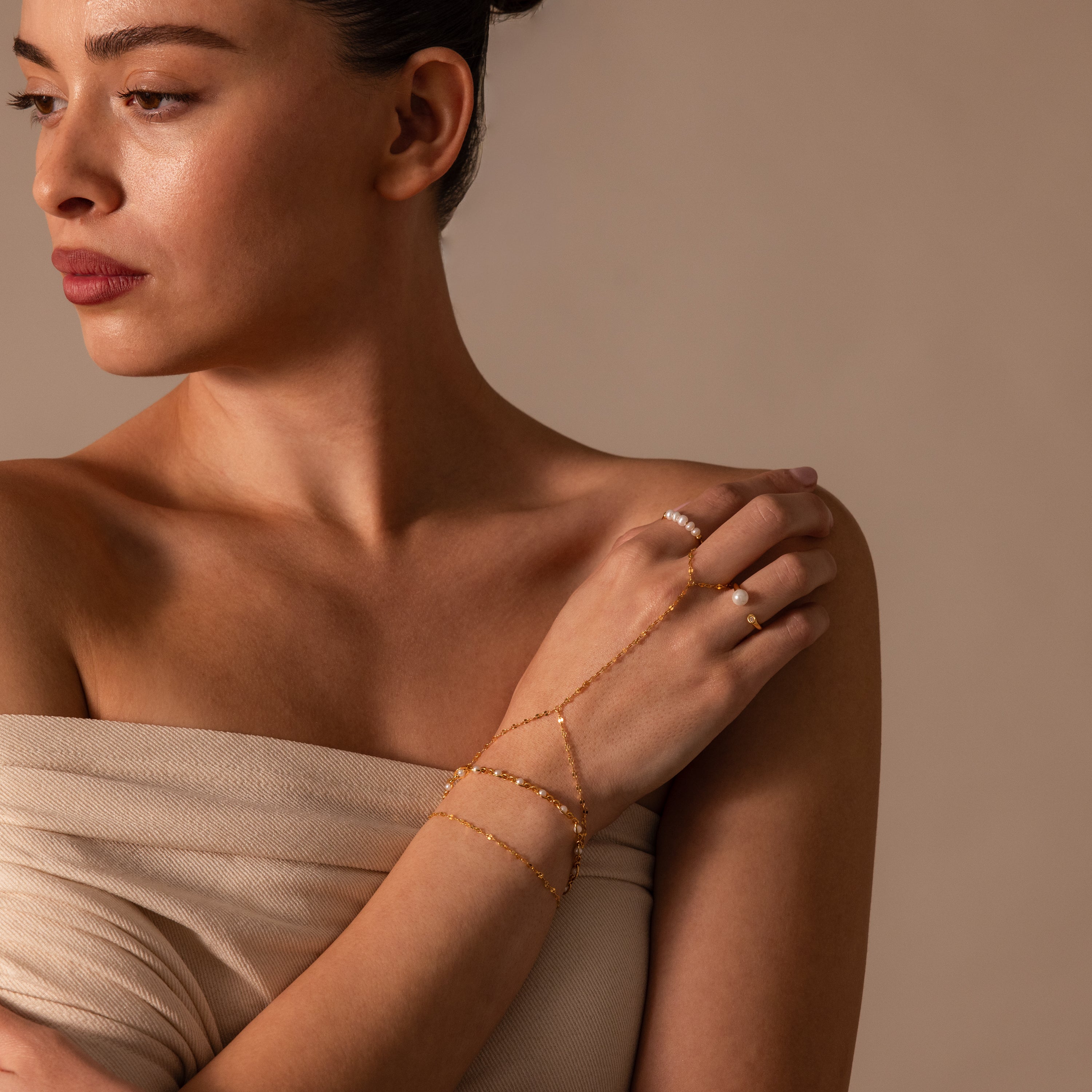 A woman in a beige dress looks to the side against a neutral background, wearing the Sparkle Hand Chain Bracelet in 18K Gold with delicate mirror cut links.