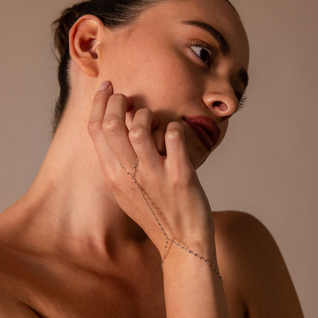 A woman with red lipstick touches her face, wearing the Sparkle Hand Chain Bracelet in Silver featuring delicate mirror cut links.