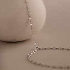 The Sparkle Hand Chain Bracelet in Silver, featuring mirror-cut links, is elegantly draped over a smooth, round beige surface on a soft beige background.
