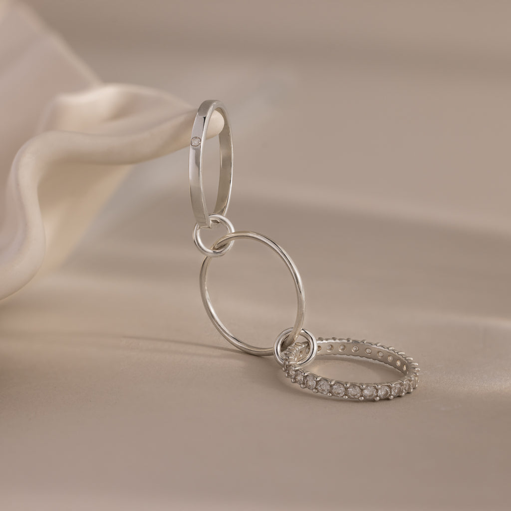 The Triple Linked Birthstone Ring Set features three silver rings, including a sparkling eternity birthstone band, elegantly linked together and displayed on a soft beige surface.
