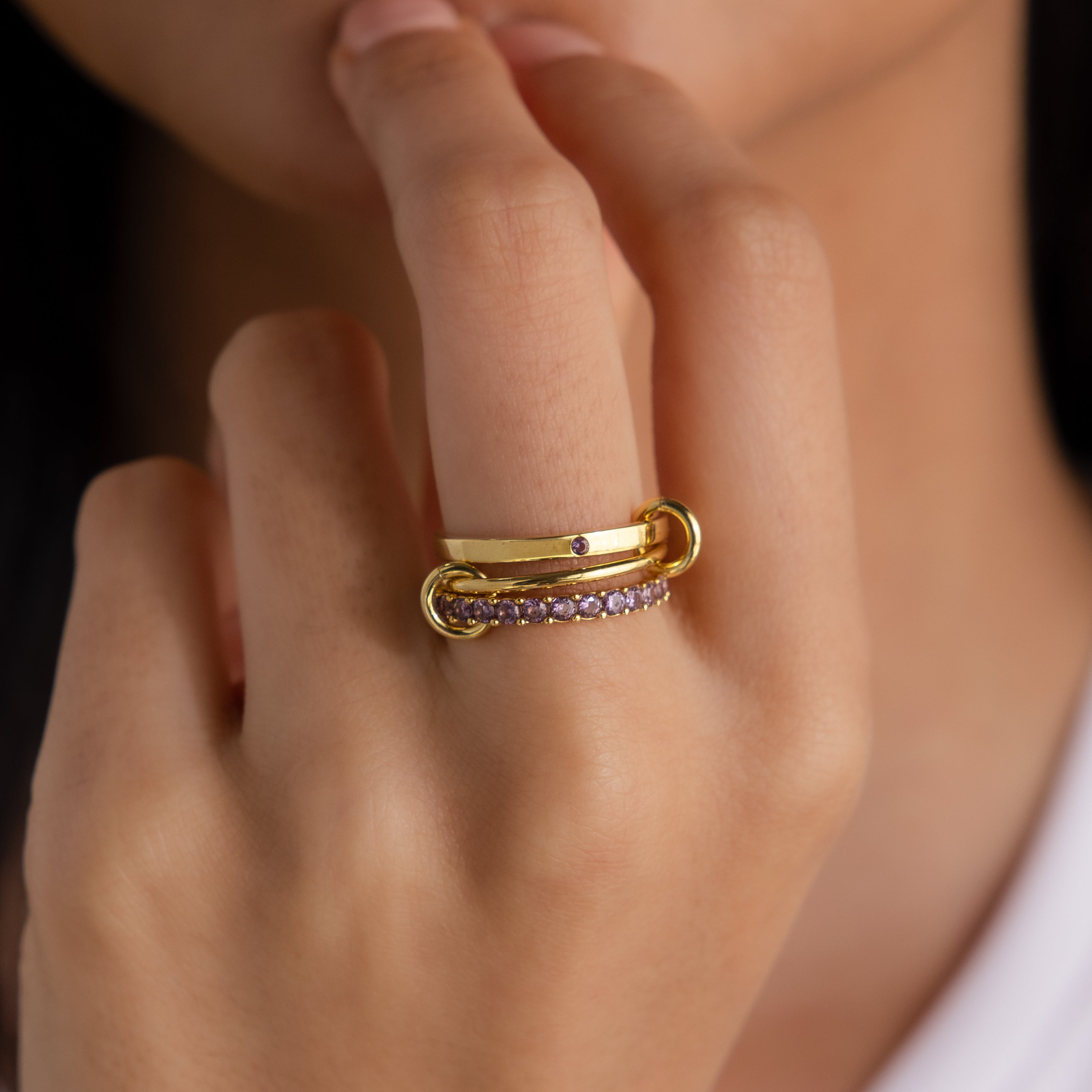 A hand with gold rings, featuring the Triple Linked Birthstone Ring Set with small purple stones, touching the chin.
