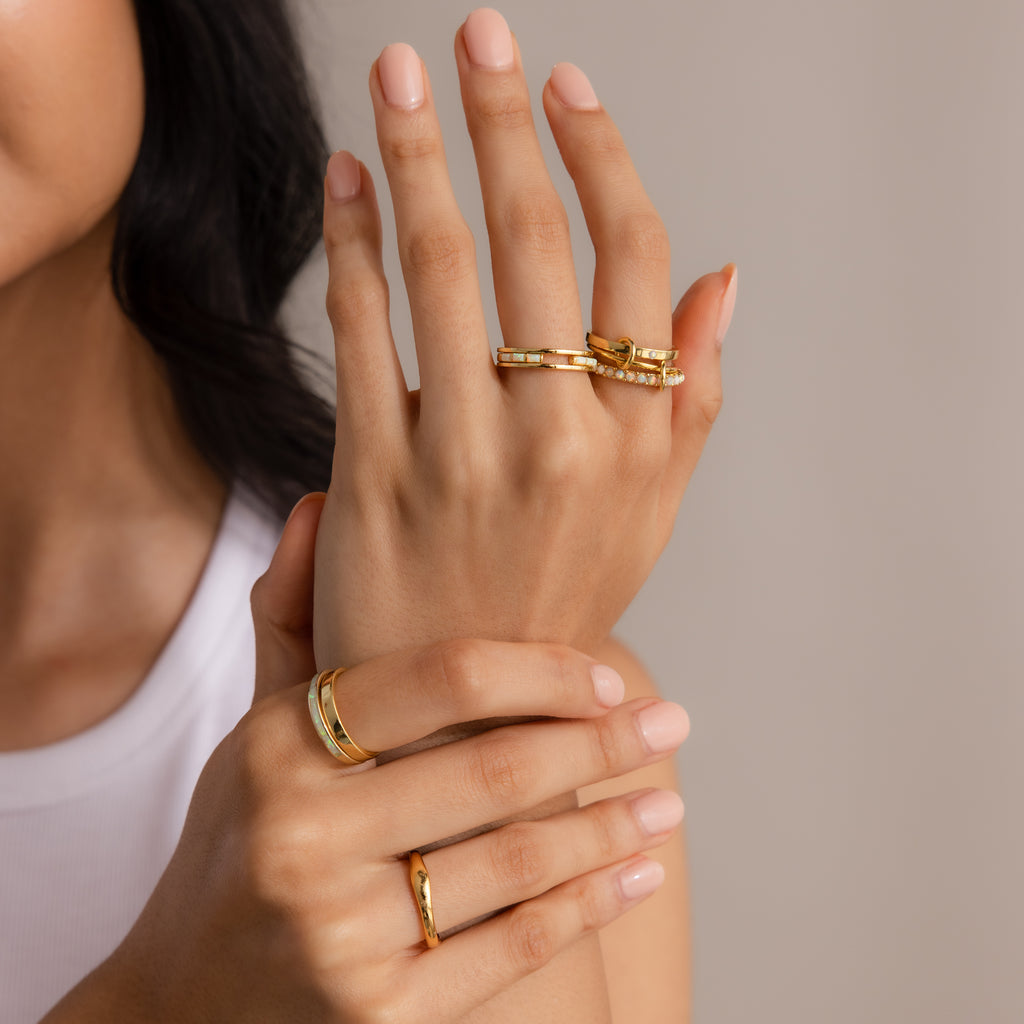 A woman's hands with nude nail polish display multiple gold rings, including the Triple Linked Birthstone Ring Set, elegantly posed against a neutral background.