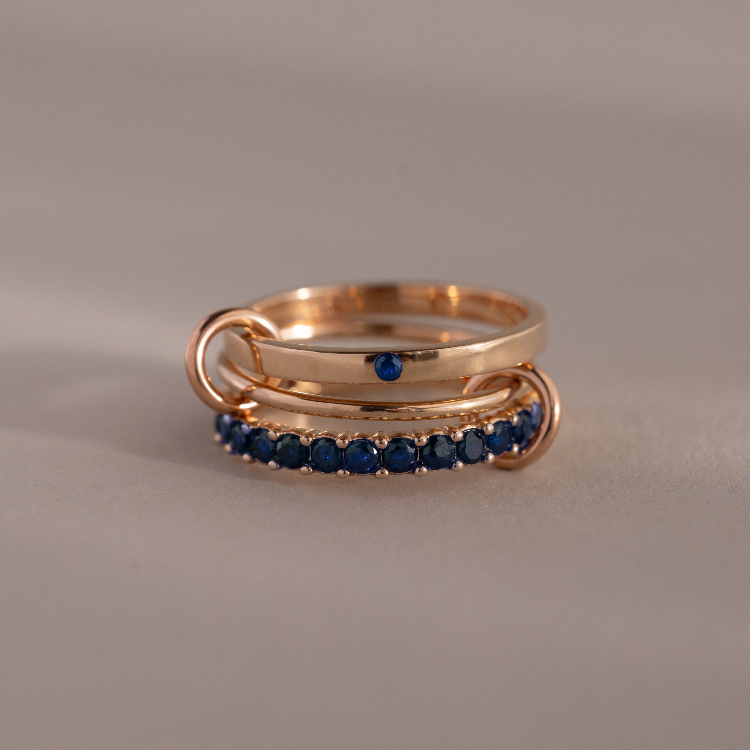 The Triple Linked Sapphire Ring Set features three intertwined gold bands—one plain, one with a single blue sapphire, and one adorned with a row of sapphires—symbolizing unity and everlasting love.